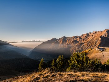 Pfunders: urig und idyllisch Das Bild zeigt eine malerische Berglandschaft bei Sonnenaufgang oder Sonnenuntergang, mit tiefen Tälern, die von Nebel bedeckt sind, und sonnenbeschienenen Berghängen, die in warmen Orangetönen leuchten. Auf einem Bergrücken rechts im Bild steht eine Berghütte, umgeben von grasbewachsenen Hügeln und Nadelbäumen.
