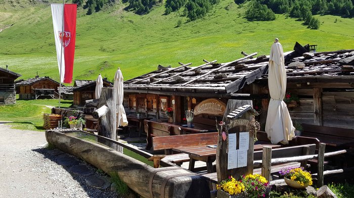 Discover the Alpine huts in Meransen, Vals & Co. The picture shows a rustic wooden hut with an outdoor area furnished with wooden tables, benches and rolled-up parasols, surrounded by a lush green Alpine landscape and a red and white flagpole.