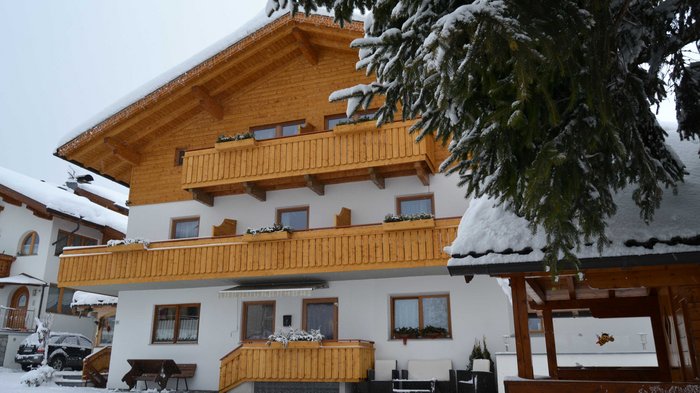 Discover the Alpine huts in Meransen, Vals & Co. A two-storey building with a white façade and wooden balconies is covered in snow. A snow-covered bench stands in front of the house and a snow-covered tree can be seen in the foreground.