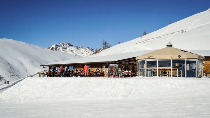 Discover the Alpine huts in Meransen, Vals & Co. The picture shows a mountain hut in the snow with a large terrace on which several people in ski clothing are sitting and relaxing. Next to the hut is a modern, glazed round building. The surroundings consist of snow-covered mountains under a clear blue sky.