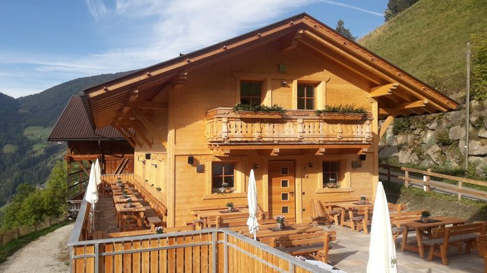Discover the Alpine huts in Meransen, Vals & Co. A mountain hut made of light-coloured wood with a balcony and flower boxes stands on a mountainside. Several wooden tables and benches with parasols are set up on a terrace in front of the hut.