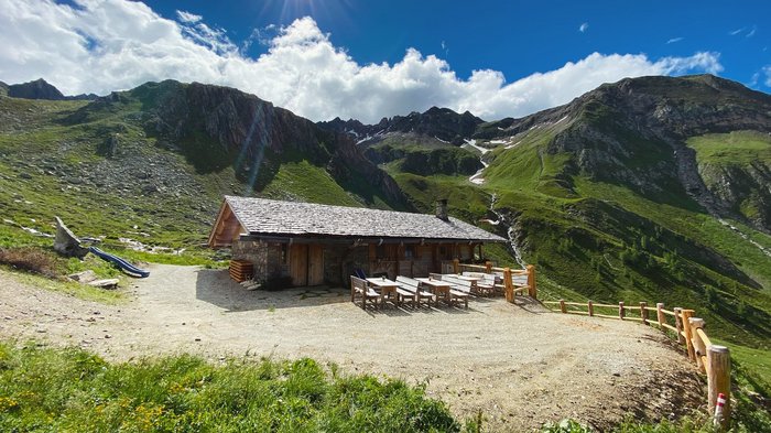 Discover the Alpine huts in Meransen, Vals & Co. A mountain hut made of wood and stone stands on a sunny, green mountain meadow, surrounded by high, rocky mountains. There are several wooden tables and benches in front of the hut.
