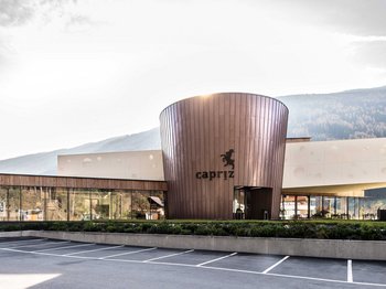 Vintl, in the Pustertal valley The picture shows a modern building with a round copper-colored facade and large glass windows. The lettering “Capriz” can be seen on the building and there is an empty parking lot in front of it, while wooded hills and mountains can be seen in the background.