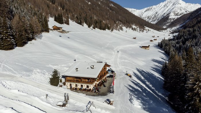 Discover the Alpine huts in Meransen, Vals & Co. A mountain hut stands in a snow-covered valley landscape, surrounded by wooded mountain slopes and other scattered huts. People can be seen in front of the hut in the foreground, while a path leads through the snow-covered landscape.