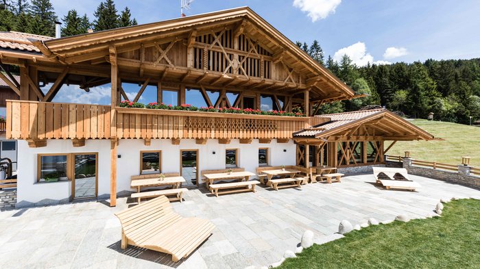Discover the Alpine huts in Meransen, Vals & Co. The picture shows an elegant, traditional Alpine house with a wooden façade and balcony, decorated with red flowers, against a backdrop of green trees and a well-kept stone slab with several wooden tables and an outdoor bench.