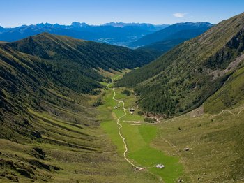 The mountain village of Meransen The picture shows a wide valley stretching between two high, wooded mountain slopes. A narrow, winding path leads through the green valley, past a few scattered huts. The majestic peaks of the Alps can be seen in the background, dominating the horizon.