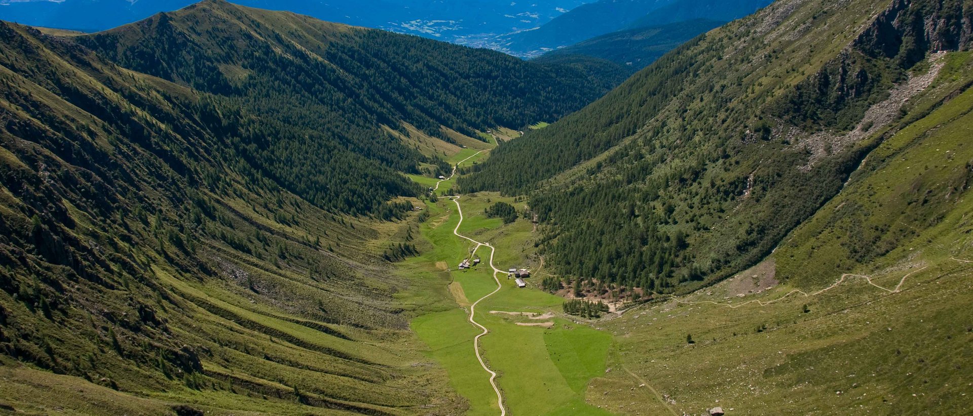 Cable Car Special – 3 in 6 The picture shows a wide valley stretching between two high, wooded mountain slopes. A narrow, winding path leads through the green valley, past a few scattered huts. The majestic peaks of the Alps can be seen in the background, dominating the horizon.