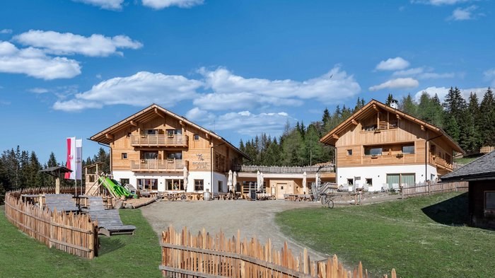 Discover the Alpine huts in Meransen, Vals & Co. The picture shows two modern wooden buildings of the "Roner Alm" under a clear blue sky with scattered white clouds. The buildings are surrounded by a wooden fence and in the foreground there is a playground with a slide and some wooden benches. Forests and hills can be seen in the background.