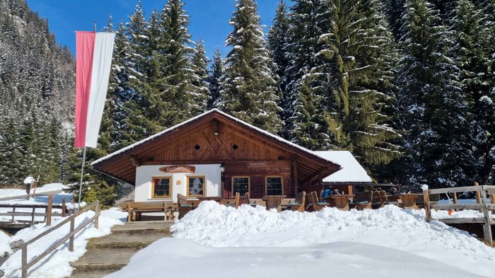 Discover the Alpine huts in Meransen, Vals & Co. The picture shows a mountain hut with a wooden façade and an Austrian flag flying in front of it. The hut is surrounded by deep snow and in the background are snow-covered conifers under a clear, blue sky.
