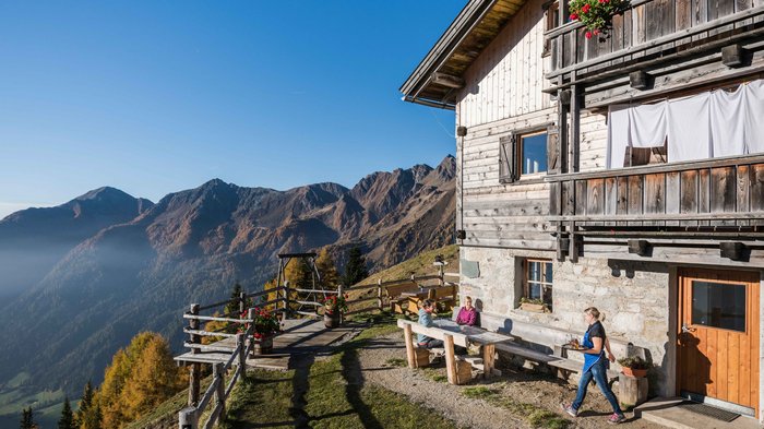 Unterkünfte Gitschberg Jochtal: Angebote Das Bild zeigt eine Berghütte, die sich auf einem sonnigen Hang befindet. Vor der Hütte sitzen Menschen an einem Holztisch, und im Hintergrund erstreckt sich eine beeindruckende Berglandschaft unter einem strahlend blauen Himmel.