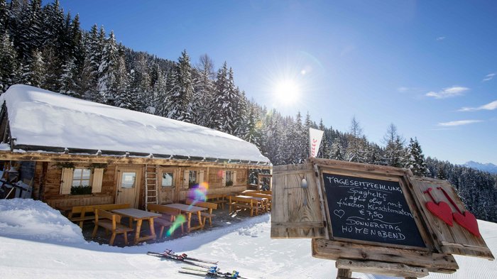 Discover the Alpine huts in Meransen, Vals & Co. The picture shows a snow-covered mountain hut with wooden benches and tables in front of the hut; next to it is a board displaying the day's recommendation, while snow-covered conifers and a bright blue sky can be seen in the background.