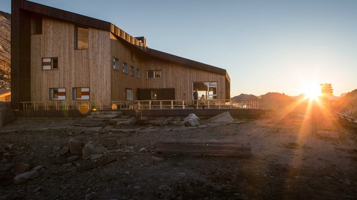 Discover the Alpine huts in Meransen, Vals & Co. The picture shows a modern wooden mountain hut in the mountains at sunset. The hut has large windows and a terrace, while the sun sets behind the mountains and bathes the surroundings in a warm light.