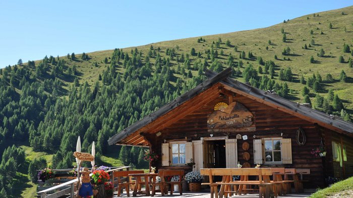 Discover the Alpine huts in Meransen, Vals & Co. The picture shows a cozy wooden mountain hut surrounded by green meadows and many fir trees. In front of the hut are wooden benches and flower boxes with colorful flowers. A wooden sign with the inscription "Welcome" greets visitors.
