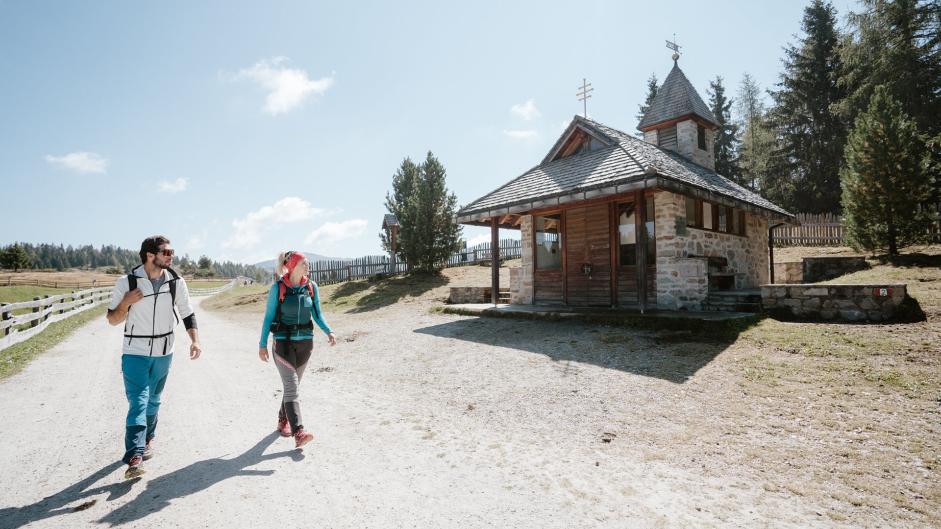 Wandelen in de Dolomieten: eenvoudigweg schitterend The picture shows two hikers, a man and a woman, walking along a wide, sandy path. To their right is a small, stone chapel with a wooden roof. The sky is clear and blue, and rolling hills and trees can be seen in the background.