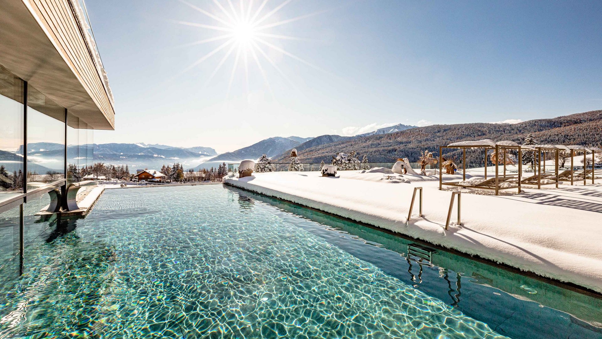 The mountain village of Meransen Infinity pool with snowy landscape in the background. The sun is shining in the sky.