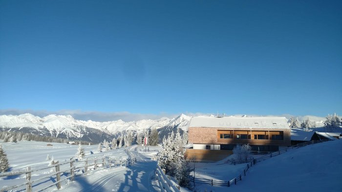 Discover the Alpine huts in Meransen, Vals & Co. The picture shows a snowy winter landscape with a modern mountain house on the right. A snow-covered path leads past the house and snow-capped mountains can be seen in the background under a clear, blue sky.