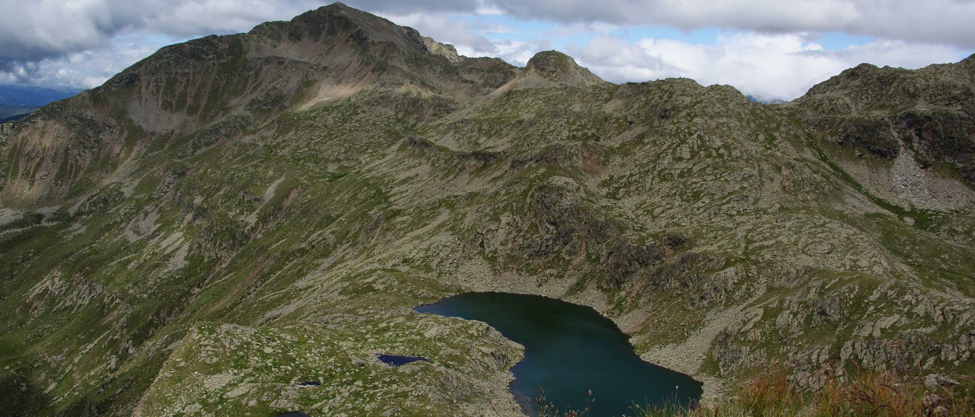 Vzhůru na Wilde Kreuzspitze! Man sieht die Eidechsspitze. Eine hügelige Landschaft im Grünen mit einem kleinen See