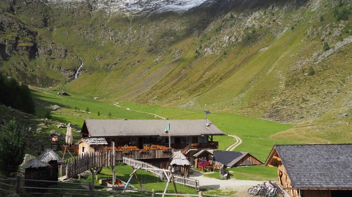 Discover the Alpine huts in Meransen, Vals & Co. The picture shows an alpine hut in a green valley surrounded by mountains. In front of the hut are wooden fences, a small bridge and several bicycles. The surrounding mountains have scattered snow peaks and a clear blue sky stretches above.