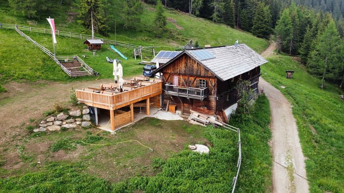 Discover the Alpine huts in Meransen, Vals & Co. The picture shows a rustic wooden mountain hut with a large terrace and balconies, surrounded by woods and meadows. There are tables and chairs in front of the hut, and solar panels can be seen on the roof.