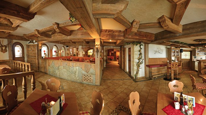 Discover the Alpine huts in Meransen, Vals & Co. The picture shows the interior of a rustic restaurant with wooden beams on the ceiling and wooden furniture. The tables are set with red placemats and traditional decorations such as deer antlers and plants hang on the walls.