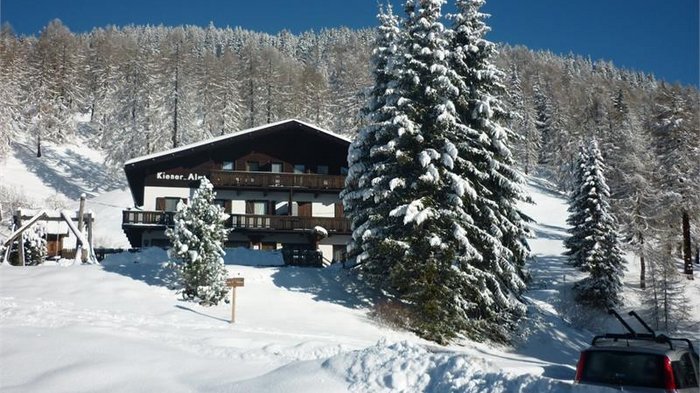 Discover the Alpine huts in Meransen, Vals & Co. The picture shows a snowy winter landscape with an alpine-style hut surrounded by snow-covered trees. A car is parked in the snow and a dense forest can be seen on a hill in the background.